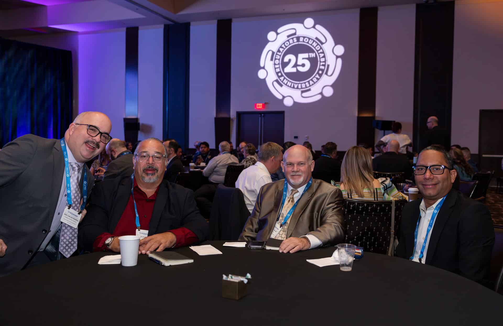 Four attendees, including Jose Carlos Figueroa, sit at a table in the event hall at the Regulators Roundtable.