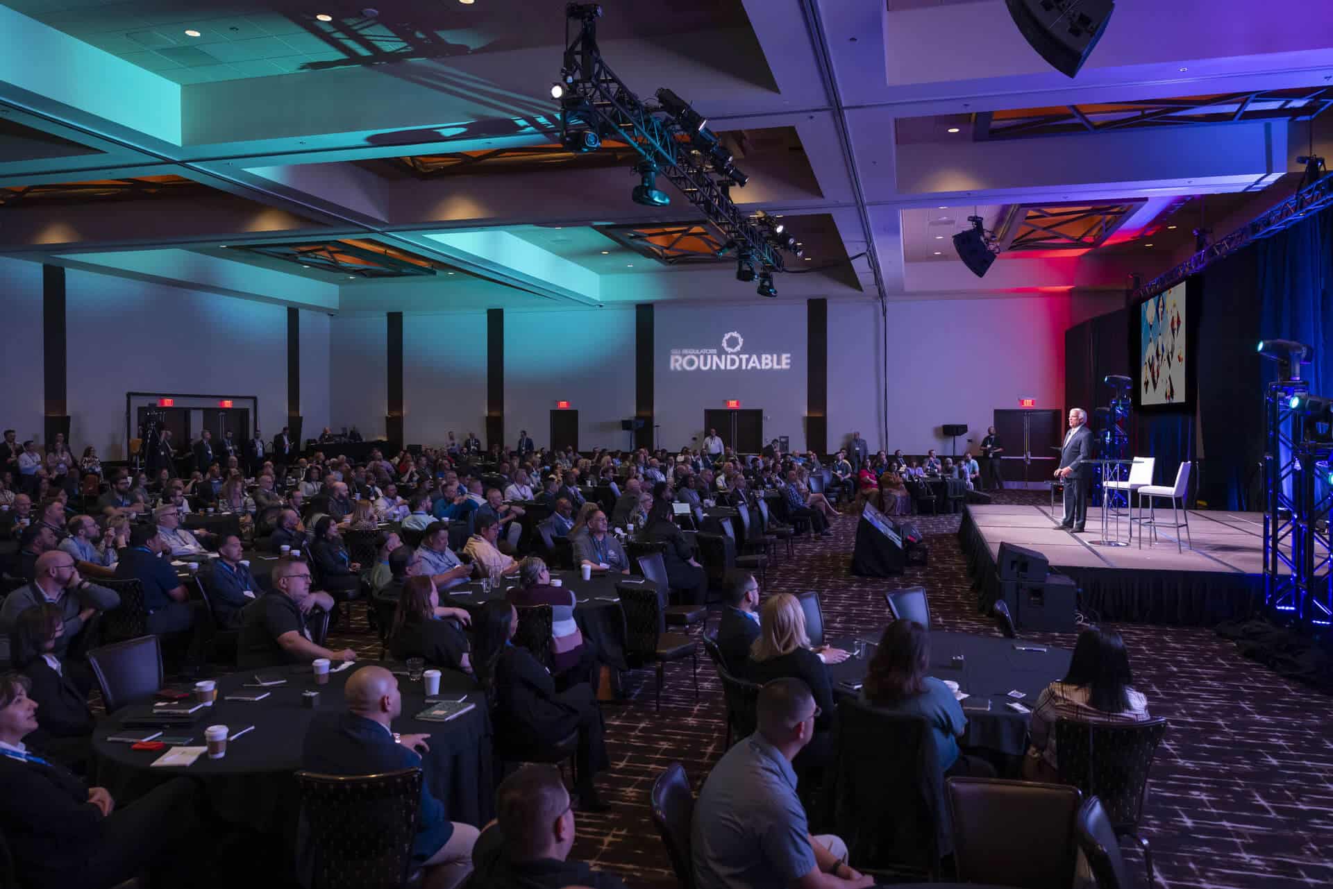 In a conference event hall, a large crowd listens intently to GLI co-founder and President James Maida presenting at the Regulators Roundtable.