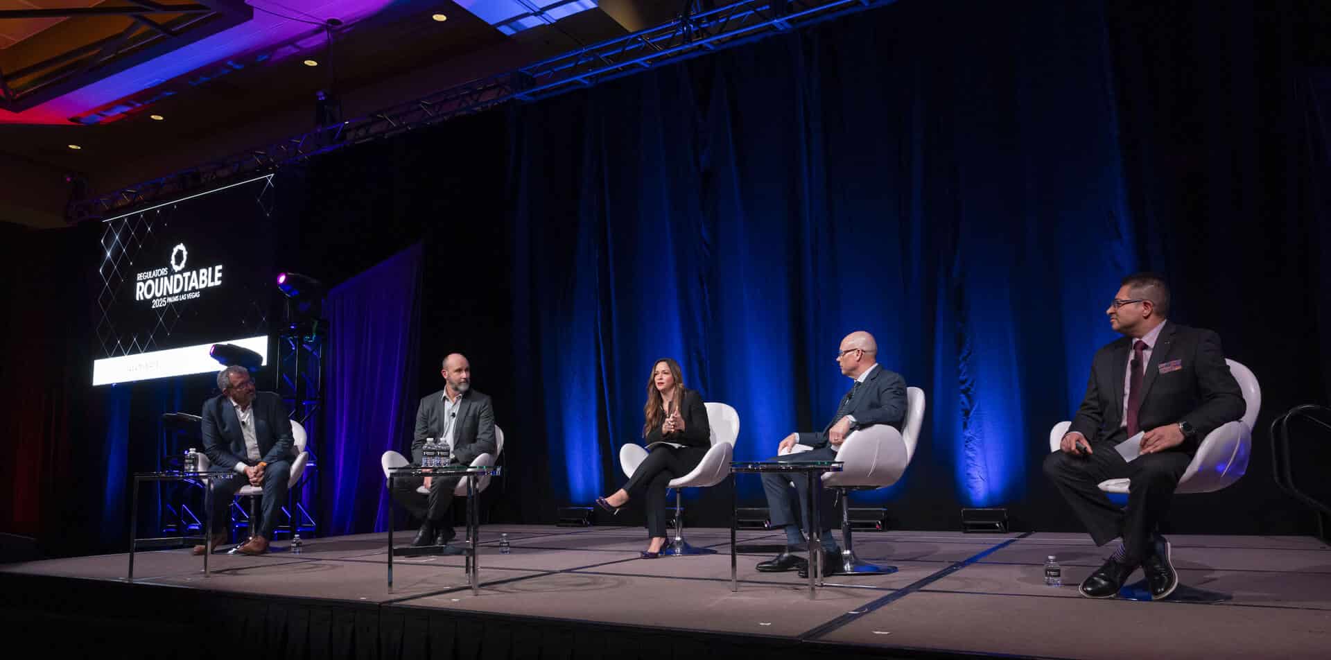 A wide view of five panelists seated on stage during a moderated discussion at the Regulators Roundtable, with a large screen and blue lighting behind them.