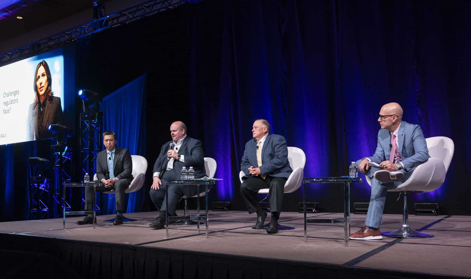 A wide view of four panelists seated on stage during a moderated discussion at the Regulators Roundtable, with a large screen and blue lighting behind them.