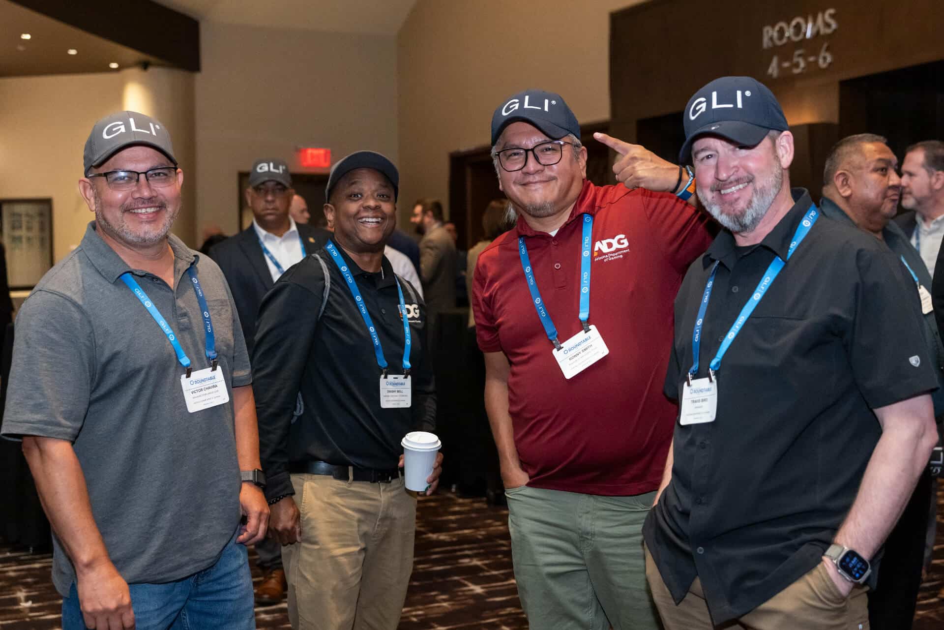 Four conference attendees wearing GLI hats and badge stand together in a busy networking area.
