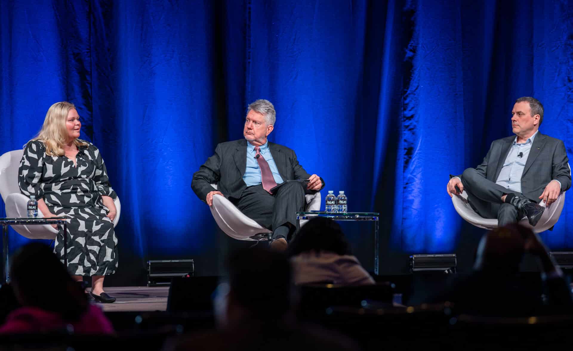 Multiple panelists participate in a formal conference session on stage during the Regulators Roundtable, seated in white chairs under professional lighting.