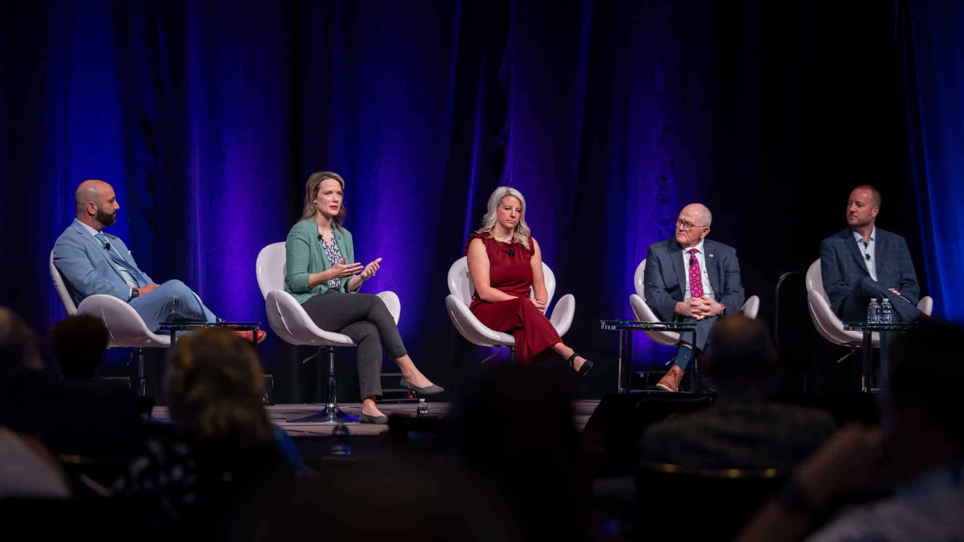 Multiple panelists participate in a formal conference session on stage during the Regulators Roundtable, seated in white chairs under professional lighting.