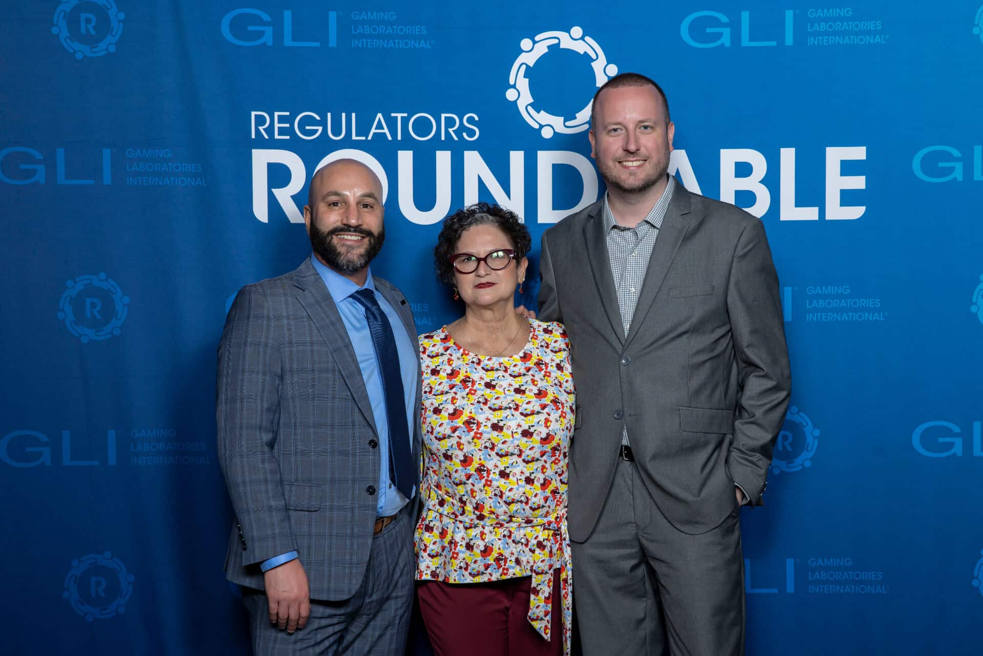 A group of three gaming professionals pose together for a photo in front of a blue Regulators Roundtable backdrop with GLI logos interpolated throughout.
