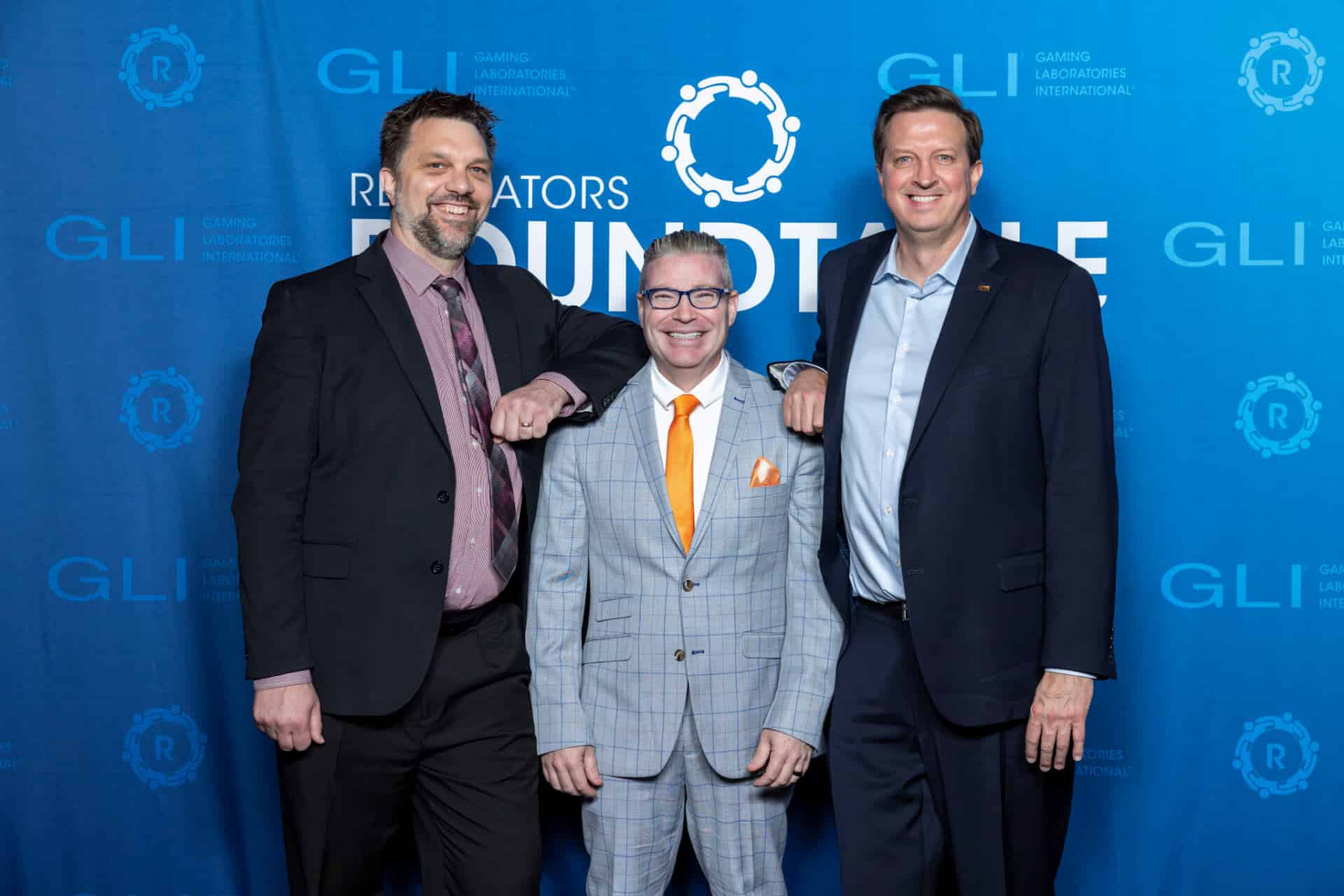 A group of three gaming professionals pose together for a photo in front of a blue Regulators Roundtable backdrop with GLI logos interpolated throughout.